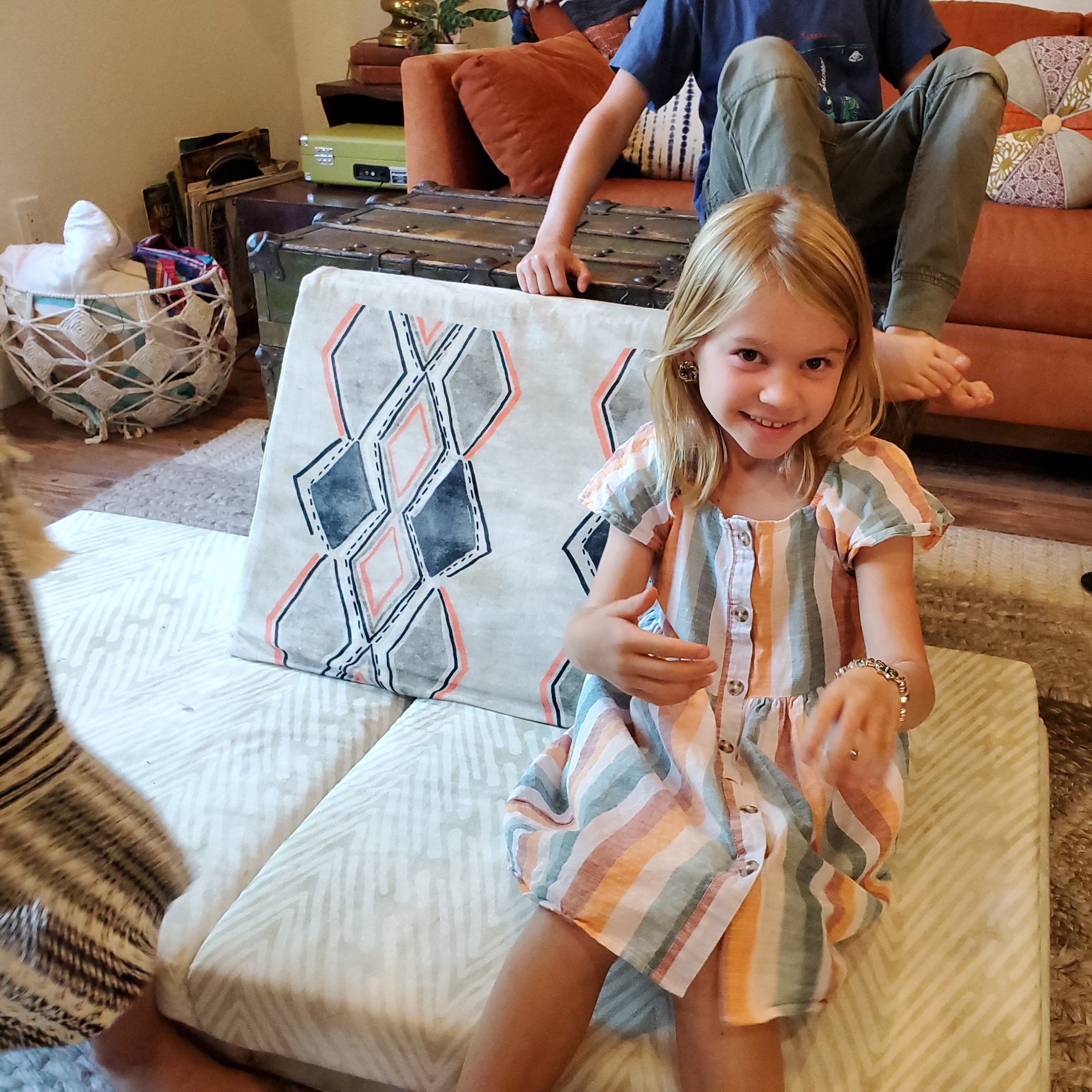 Young girl sitting on a couch holding a pillow with a geometric pattern, with a person sitting on a couch in the background.